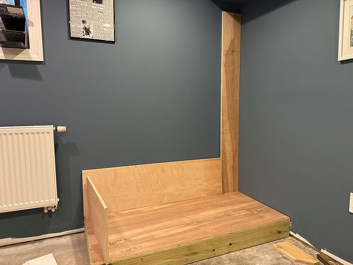 Alt text: A four-photo collage documenting the construction of a custom built-in reading nook. The top-left image shows an empty corner of a room with blue walls, white trim, and a ceiling bulkhead. The top-right image shows the initial framework for a bench seat and a vertical cabinet side installed beneath the bulkhead. The bottom-left image shows the bench structure further along, with additional framing and support panels in place. The bottom-right image shows the nearly completed project, including a built-in bench with storage compartments below and a floor-to-ceiling bookshelf fitted beneath the bulkhead. Together, the images illustrate the progression from an empty corner to a custom built-in seating and storage feature.