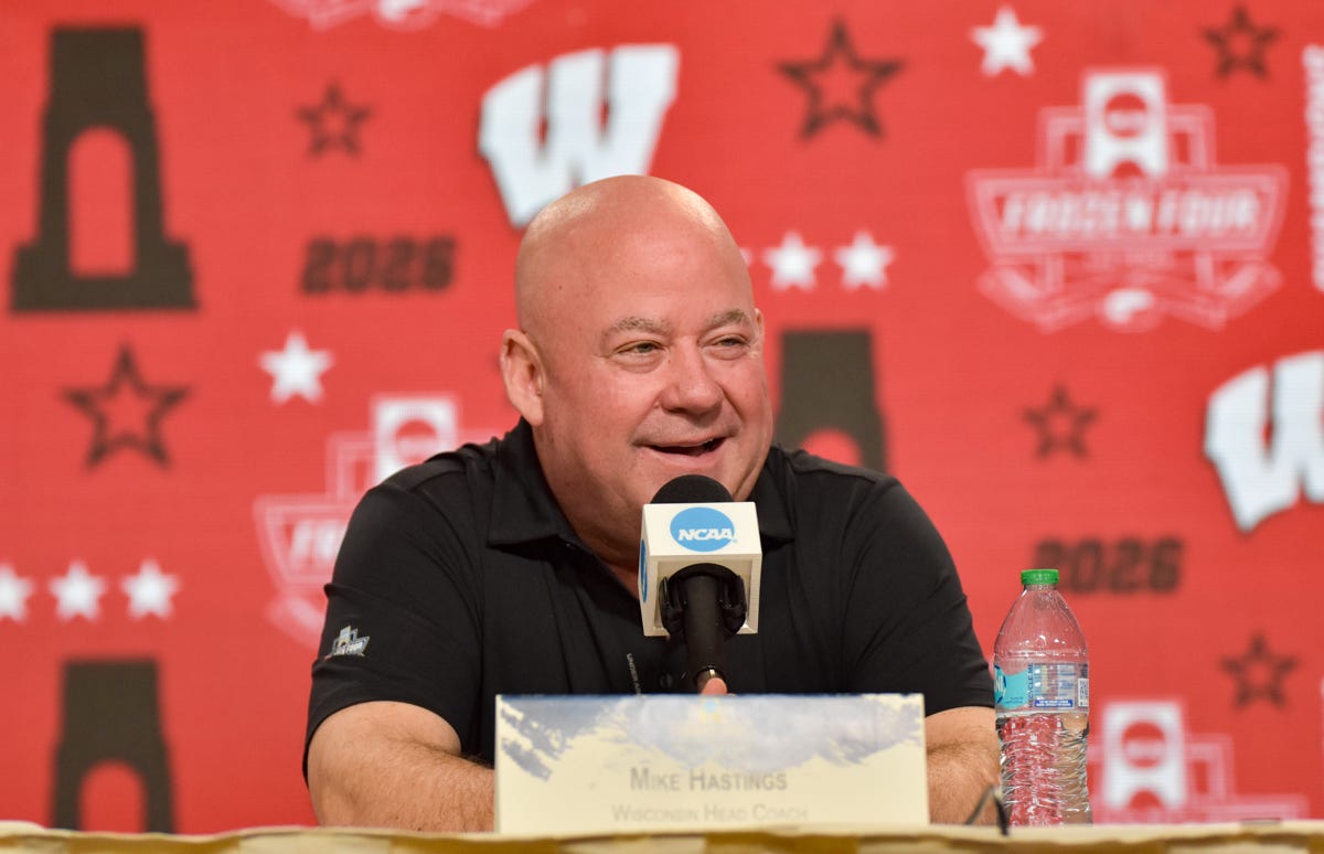Mike Hastings sits at a podium in front of a Wisconsin Badgers Frozen Four backdrop
