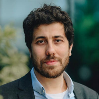 Retrato de um homem jovem de cabelos castanho-escuros ondulados usando camisa azul e blazer. Olha para a câmera com leve sorriso, diante de um fundo vegetal desfocado. Retrato de um homem jovem de cabelos castanho-escuros ondulados usando camisa azul e blazer. Olha para a câmera com leve sorriso, diante de um fundo vegetal desfocado.