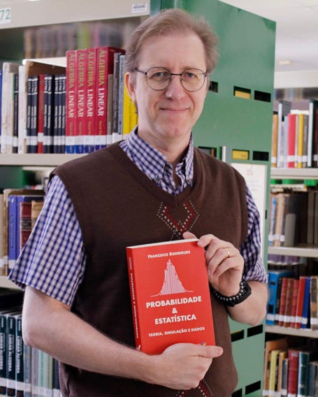 Fotografia de um homem em pé em uma biblioteca, entre estantes repletas de livros. Ele sorri discretamente para a câmera, usa óculos de armação redonda, camisa xadrez azul e branca sob um colete marrom e segura, com as duas mãos, um livro de capa vermelha intitulado Probabilidade & Estatística: Teoria, Simulação e Dados.