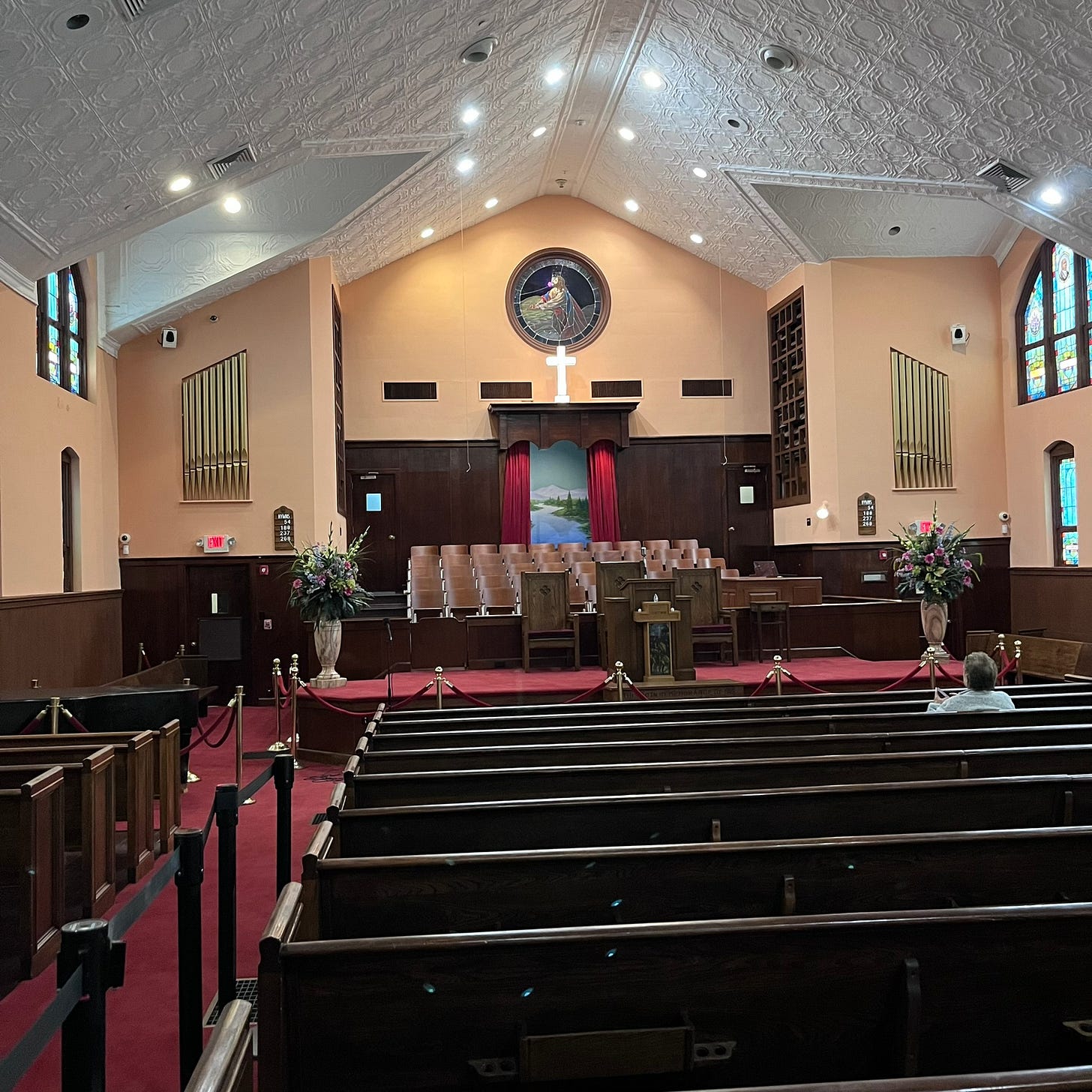photograph showing the interior of the Ebenezer Baptist Church, Atalanta. It has white appex roof, salmon pink walls, simple stain glass, an organ, stage and Baptish church iconography, it can seat several hundred parisheners, the photo is take from the back of the church looking towards the stage. One person sits by themsleves where the congregation would sit