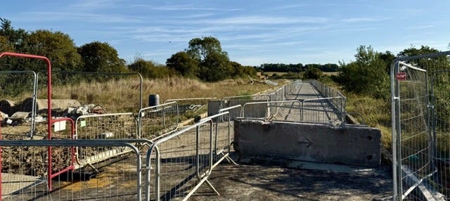 metal barriers and a large concrete slab, indicating closure or construction. The path is surrounded by grassy fields and scattered trees under a clear blue sky. No vehicles or people are present.
