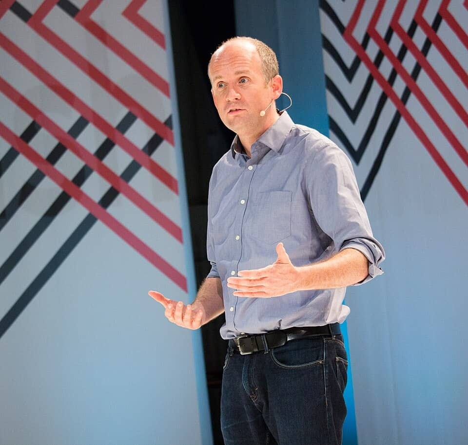 A speaker presenting on stage, gesturing with hands, against a backdrop of angled red and black lines.