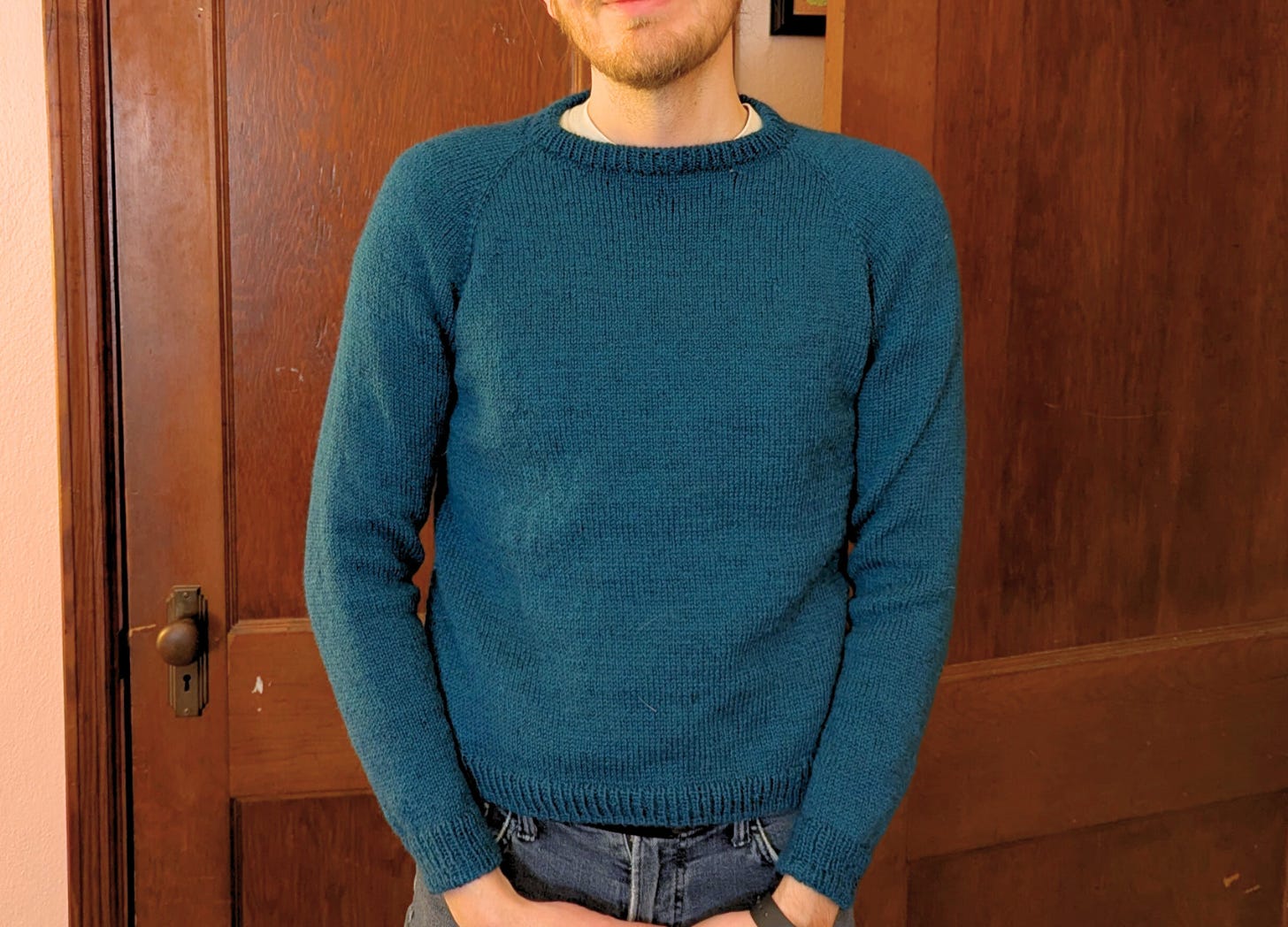 ID: A male person stands with his hands held in front of his waist. He is wearing a teal knit raglan sweater. He is standing in front of brown wooden doors. His face is obscured.