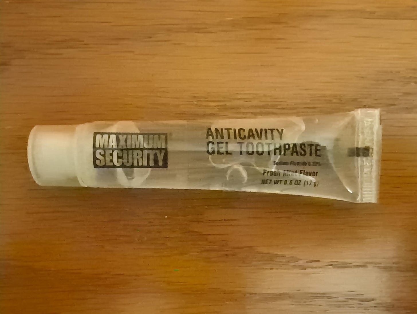 A small, nearly empty tube of 'Maximum Security' brand anticavity gel toothpaste, fresh mint flavor, 0.6 oz, lying on a wooden surface. The kind issued in jail.