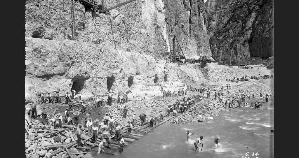 Fotografia storica in bianco e nero che mostra decine di operai al lavoro durante la costruzione della diga di Camarasa, intenti a scavare, trasportare materiali e lavorare lungo un fiume, ai piedi di un'imponente parete rocciosa.