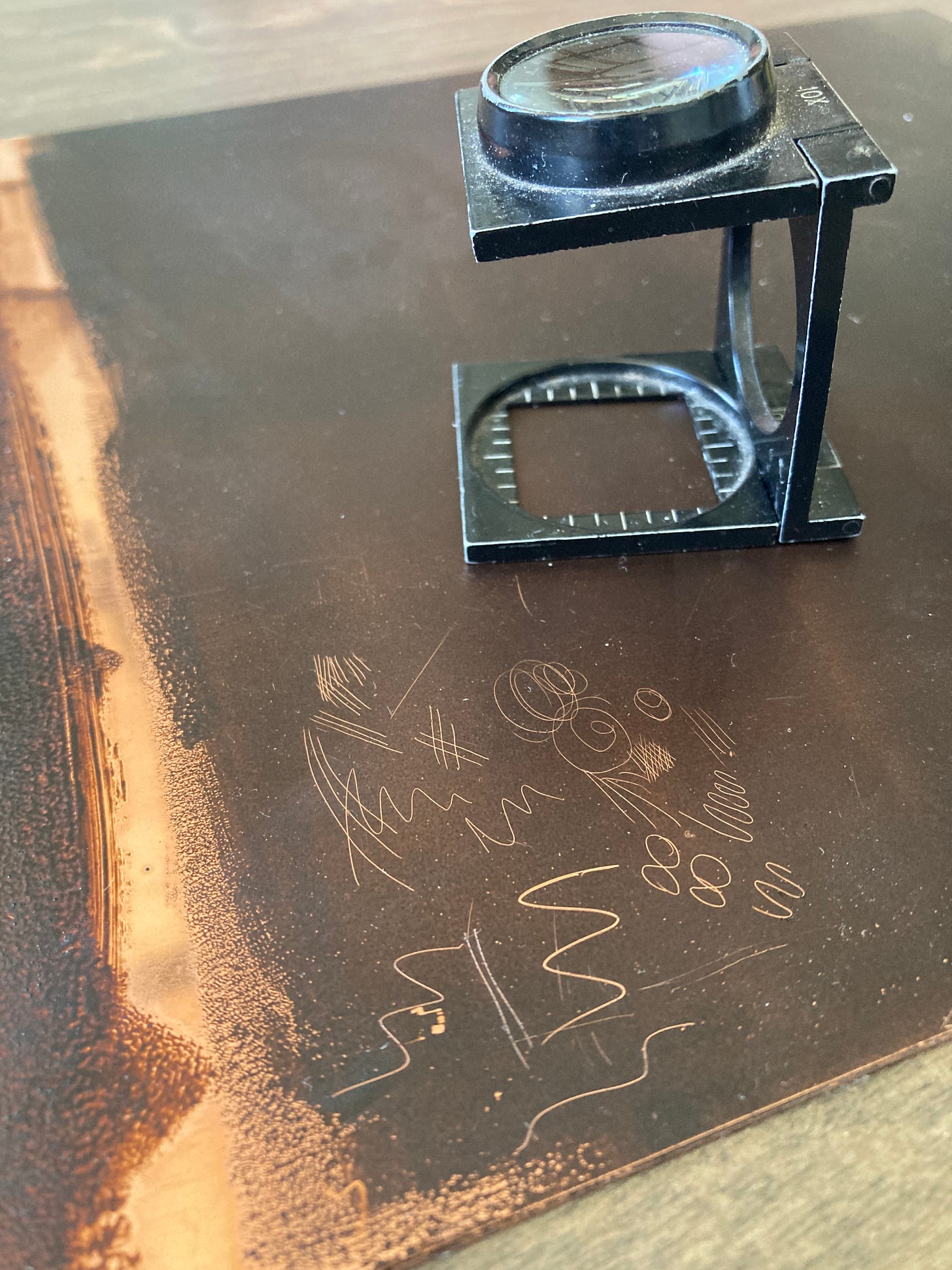 A copper plate coated with a matte brown film of hard ground fills most of the frame. Lines and scribbles have been through the ground, exposing bright copper.  A loop rests on the plate. 