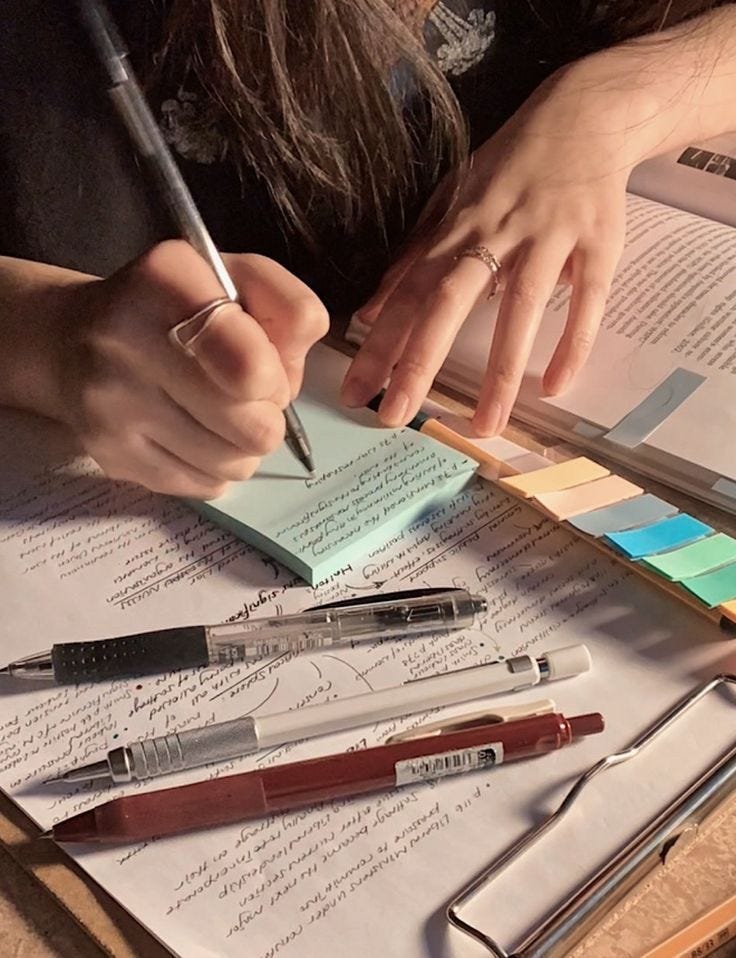 This may contain: a person is writing on some paper and holding a pen in their left hand, with several other pens sitting next to them