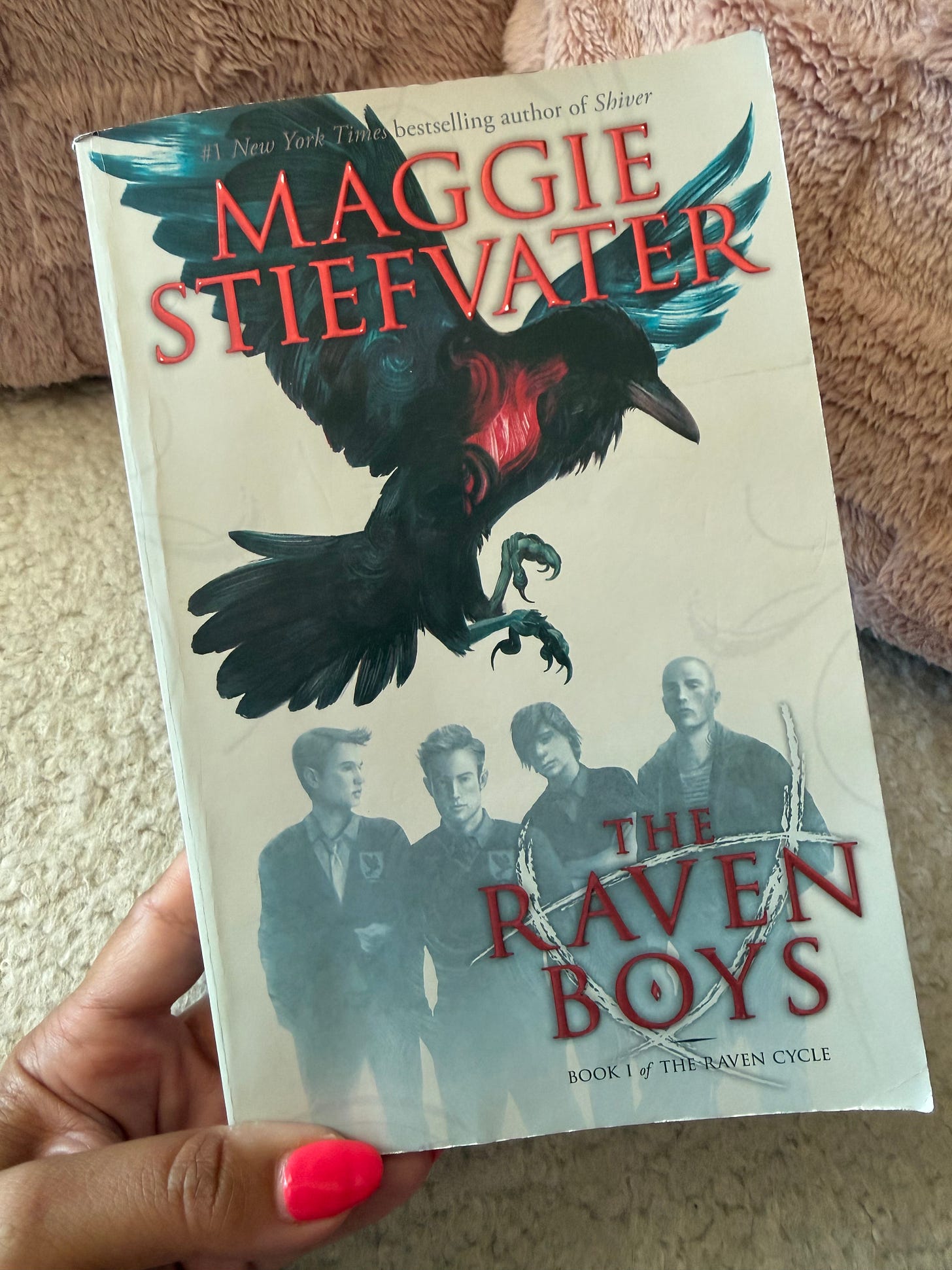 A hand holding the book cover of The Raven Boys by Maggie Stiefvater A hand holding the book cover of The Raven Boys by Maggie Stiefvater