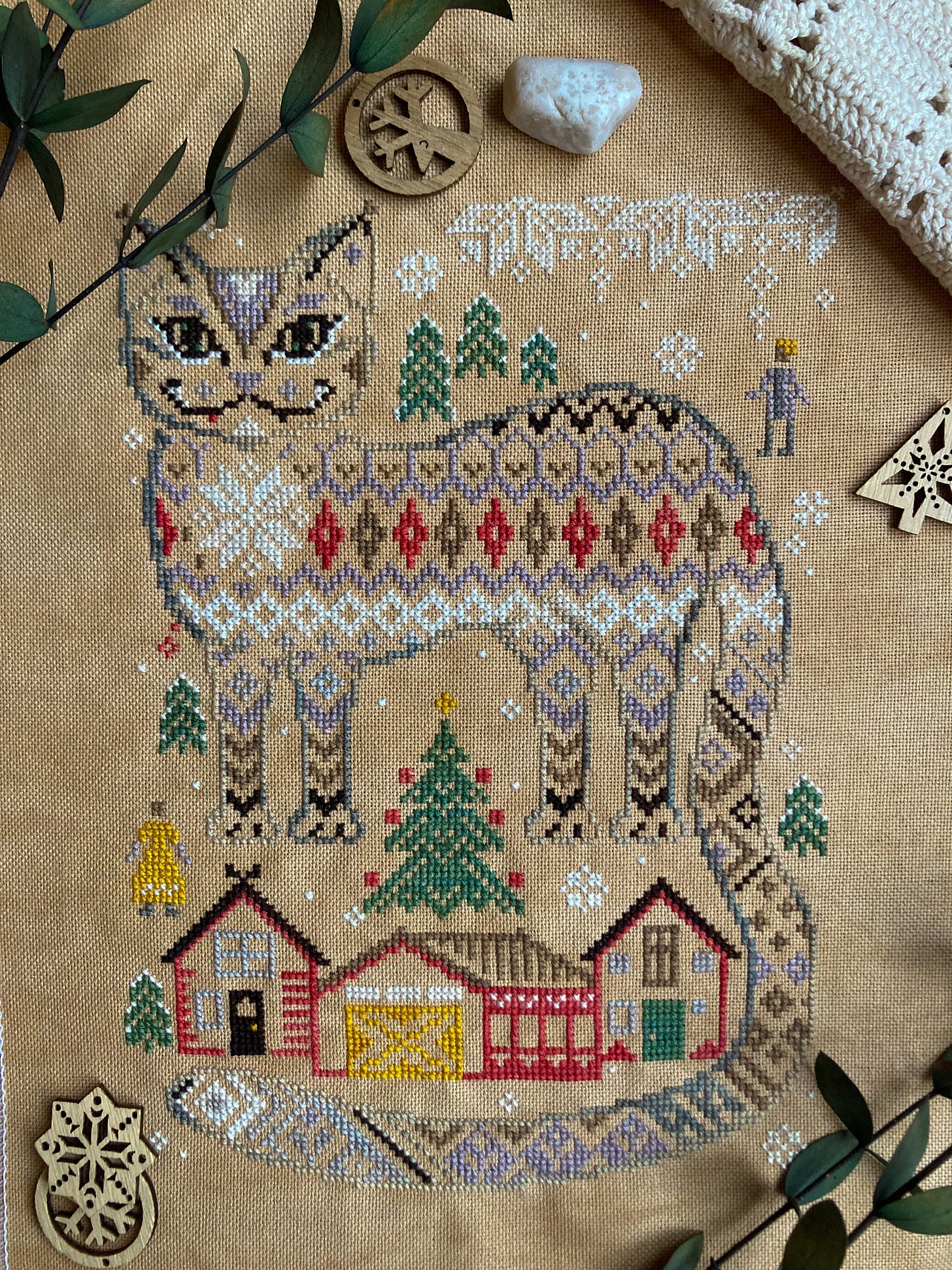 A cross stitch sampler depicting a large Yule Cat comprised of repeating patterns, looming over a small house and barn.