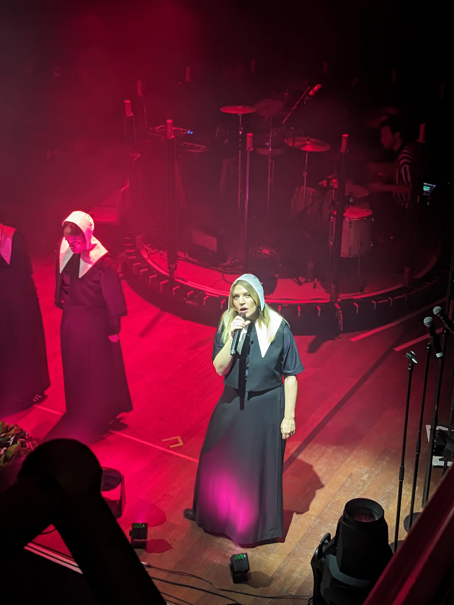 Rebecca Lucy Taylor, known as Self Esteem, is on stage with a mic singing, surrounded by other dancers. She has blonde hair and is wearing a kind-of Handmaid’s Tale-inspired outfit with a white hood. She is looking up, as I took the photo from the balcony. 