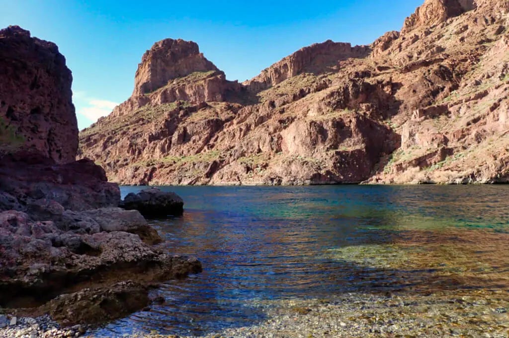 The Colorado River flows through Black Canyon beneath rugged desert cliffs. Clear blue water laps against a rocky shoreline under a cloudless sky.