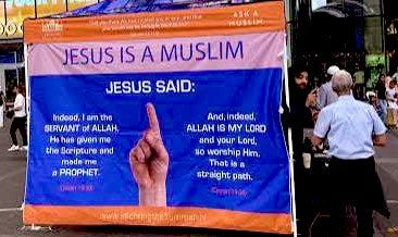 Large blue and orange tent setup at outdoor event displays bold text Jesus is a Muslim with Bible quote from John 5:30 about being servant of Allah and prophet, surrounded by diverse group of people including men in casual attire standing nearby under sunny sky.