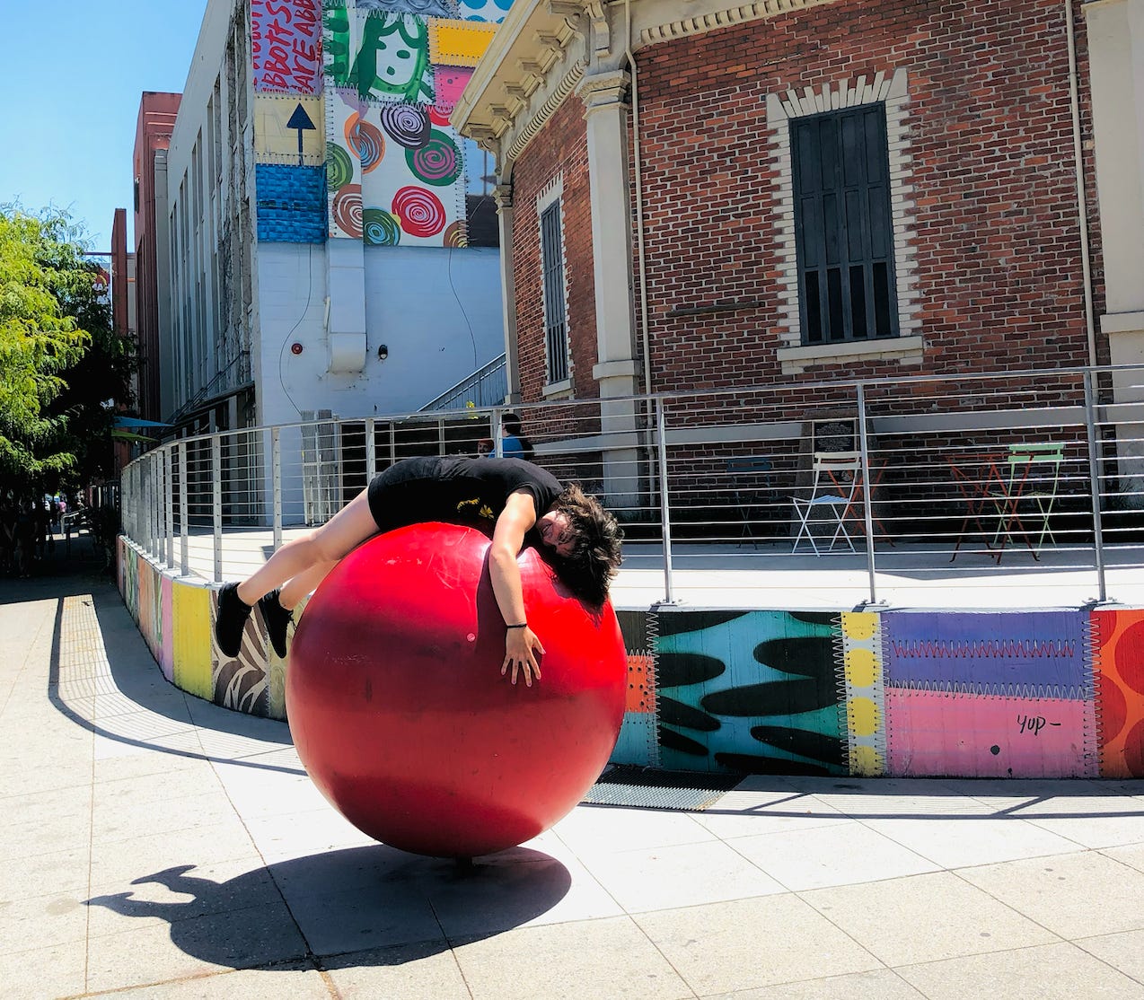 Photo of a white woman in black shirt and shorts hugging a large red ball sculpture outside of a mural and museum