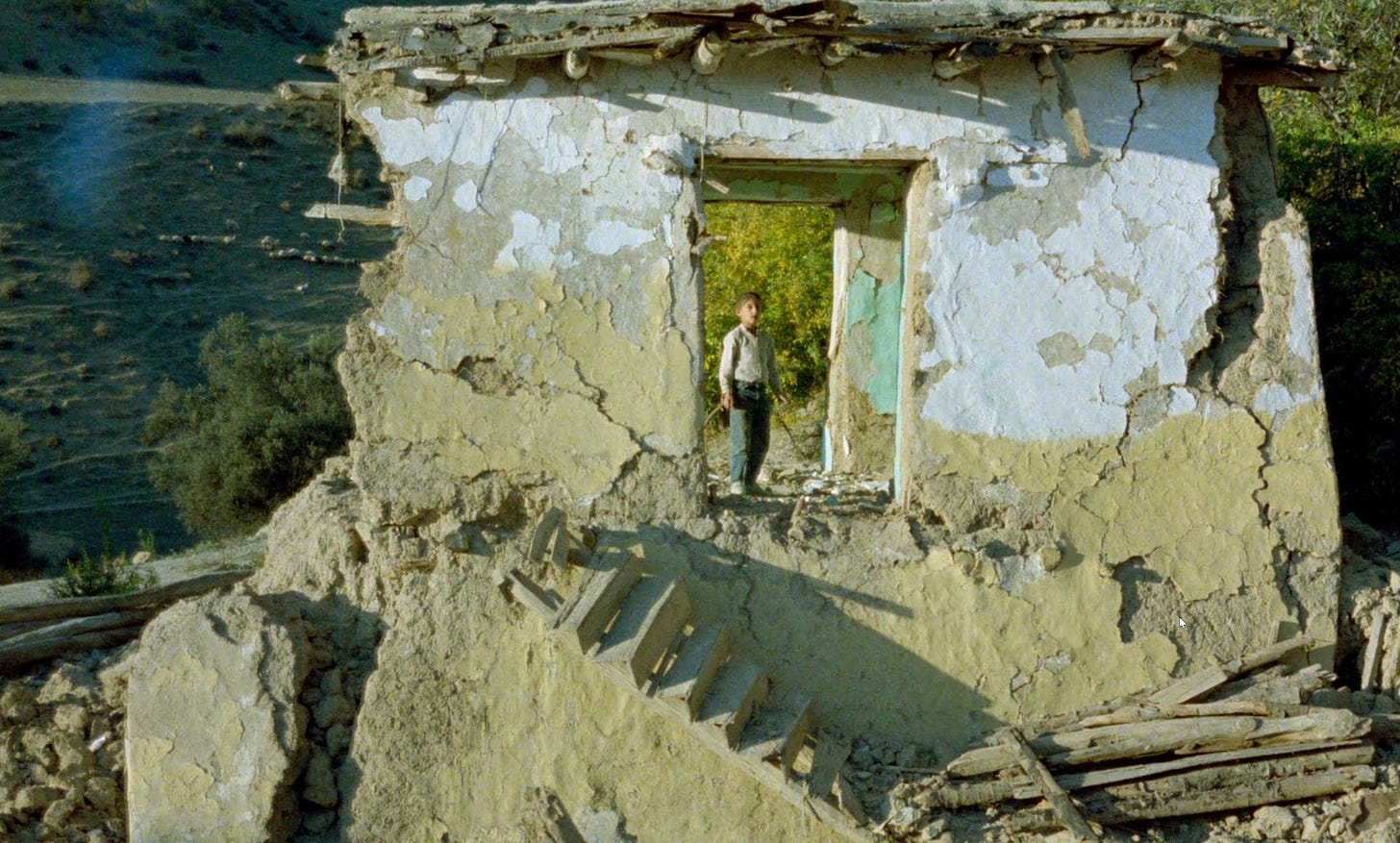captura de tela do filme "e a vida continua", de abbas kiarostami. imagem de uma casa severamente comprometida pelo terremoto, com uma parte da fachada ainda de pé, onde está o filho do diretor. ele usa uma camisa azul e uma calça jeans, e está olhando em direção à câmera, filmado de longe.