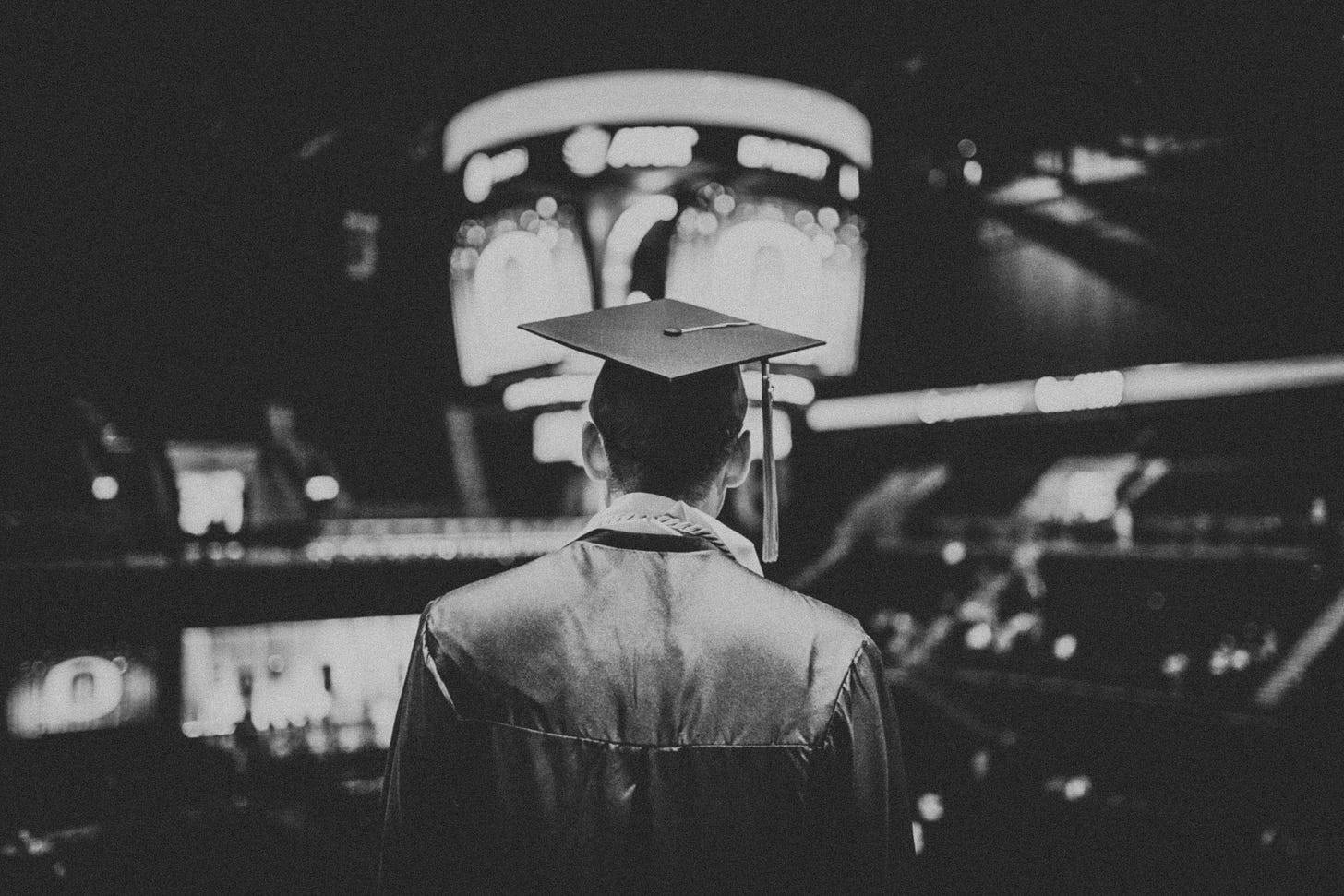 Black and white image of the back of a man wearing graduation cap and gown in an auditorium