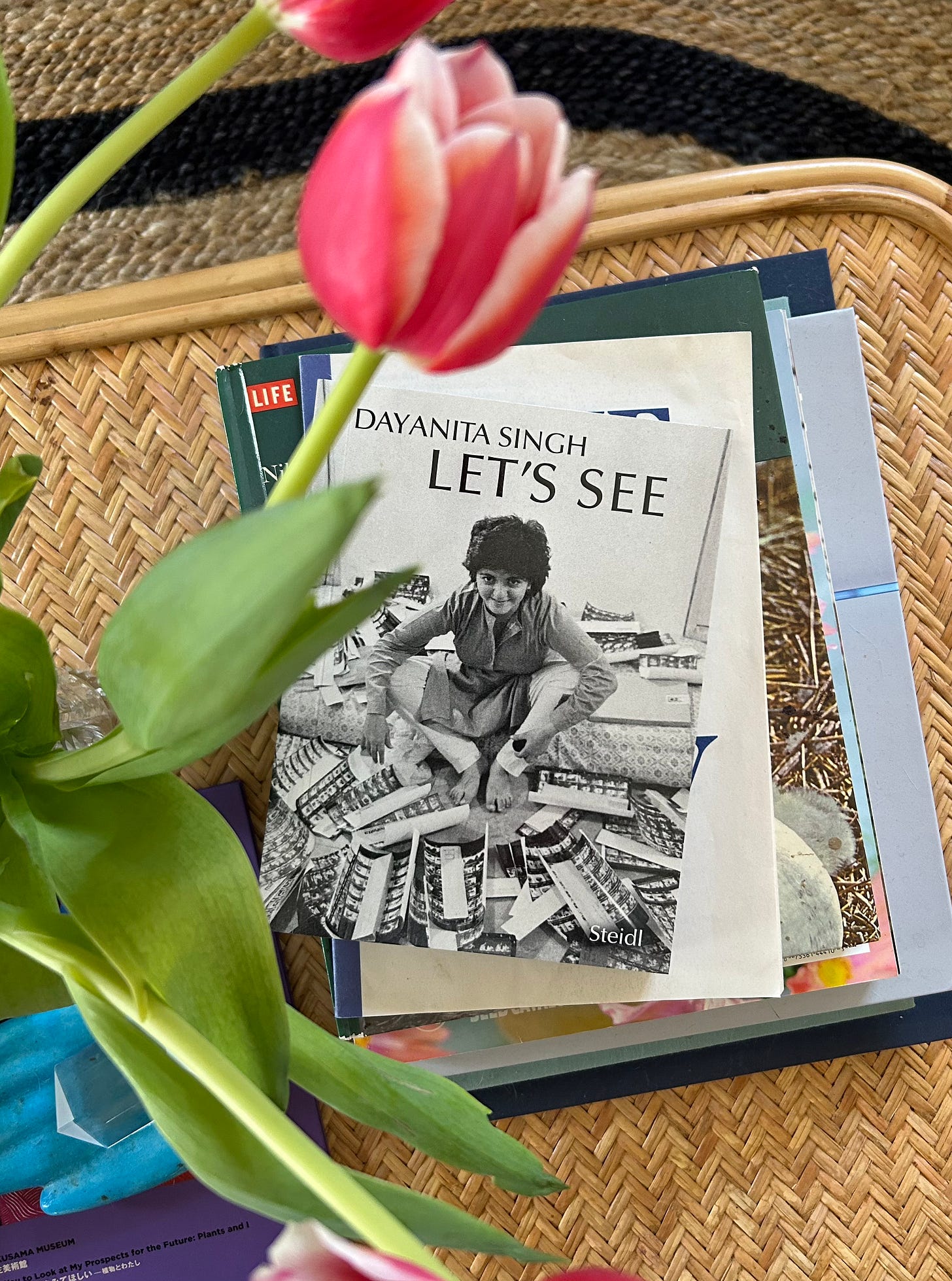 a large pink tulip takes up the top left of the image with light green leaves! Underneath that on a stack of books that are mostly gray and cream and blue is a book called Let's See by Dayanita Singh, its black and white and on the cover is Dayanita sitting around negatives of photos and pieces of paper. underneath the books is a light tan coffee table with a chevron pattern!