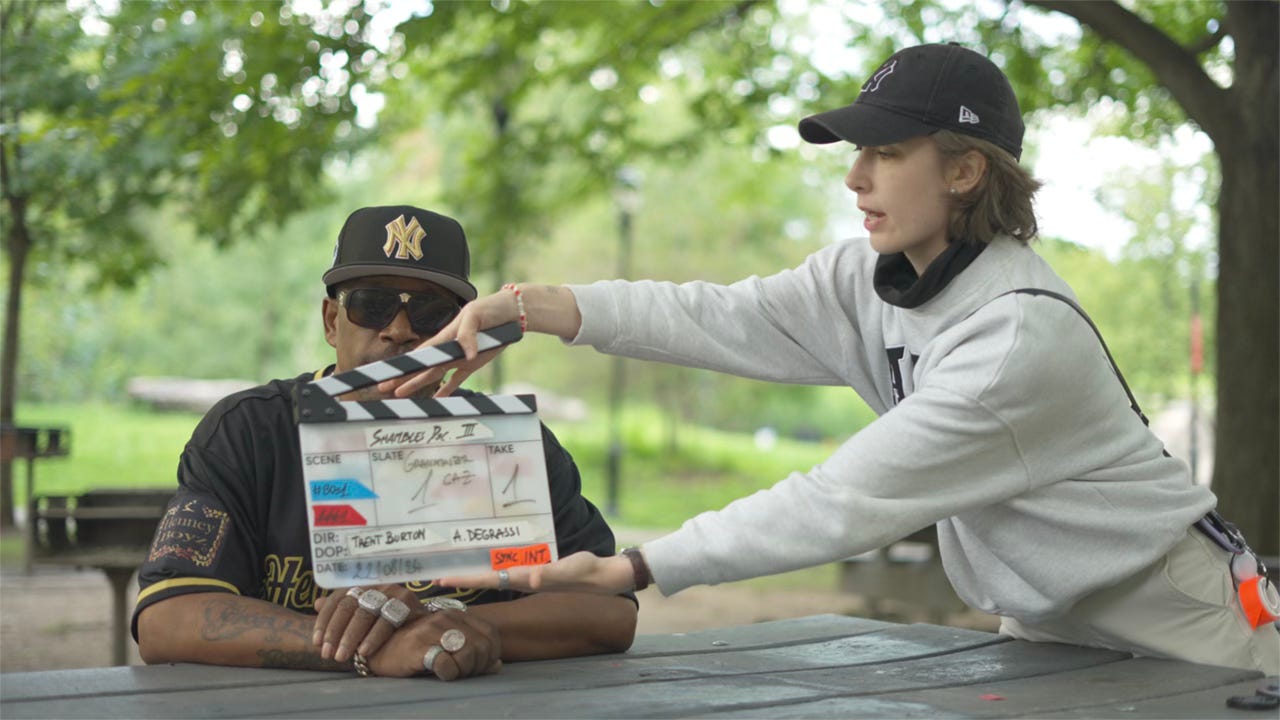 A woman holds a clapperboard in front of a man wearing a black and gold Yankees hat, black and gold sports shirt and dark sunglasses. They are in a park. A woman holds a clapperboard in front of a man wearing a black and gold Yankees hat, black and gold sports shirt and dark sunglasses. They are in a park.