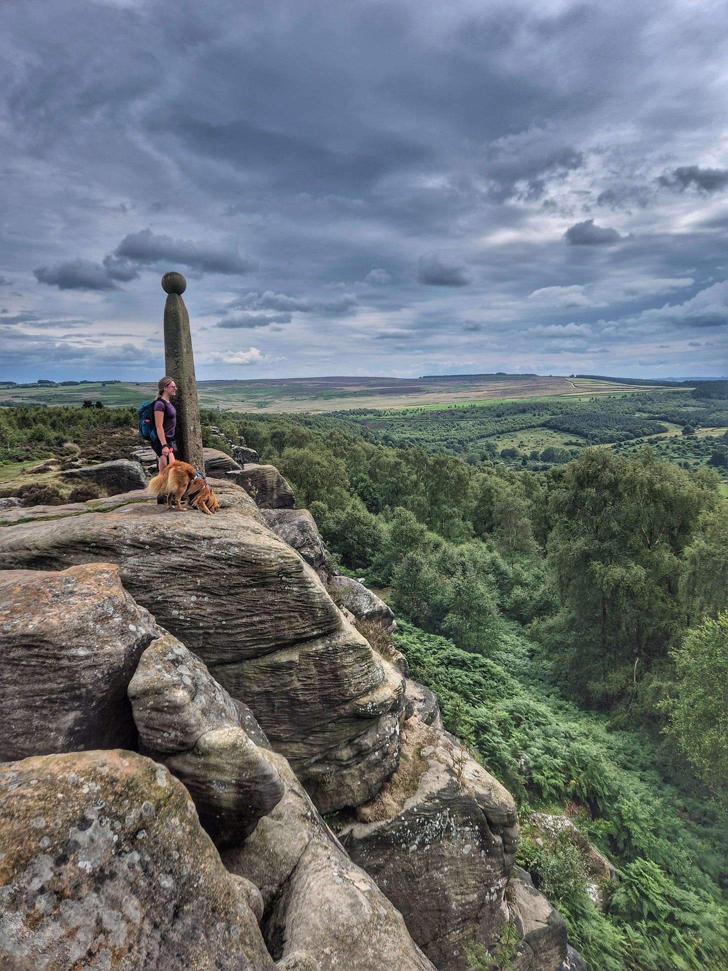 Female hiker and her dog stand at Nelson's Memorial overlooking Birchen Edge and Gardom's Edge in the distance. 