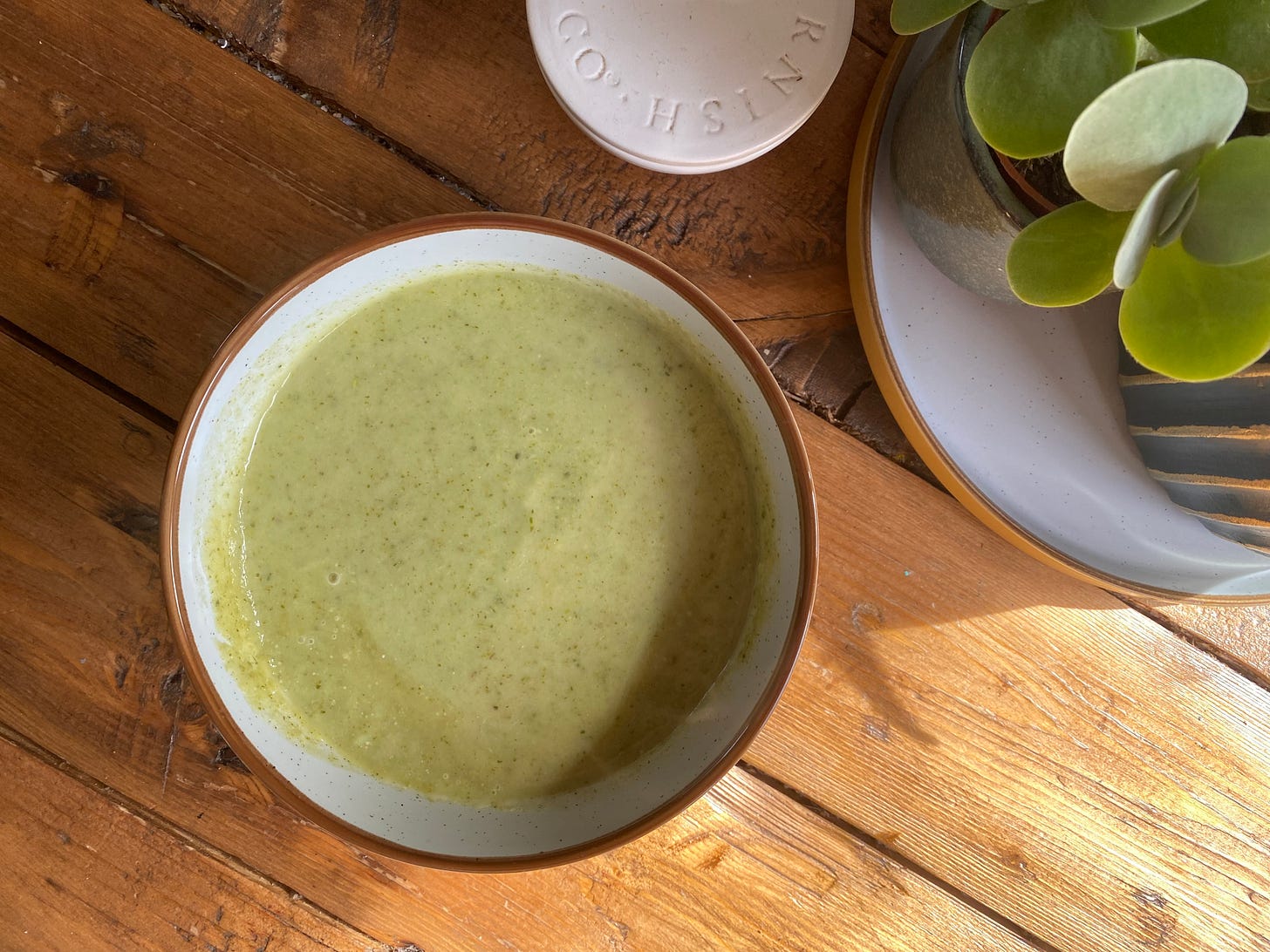 Bowl of green soup on a wooden table, next to a salt pot and house plant