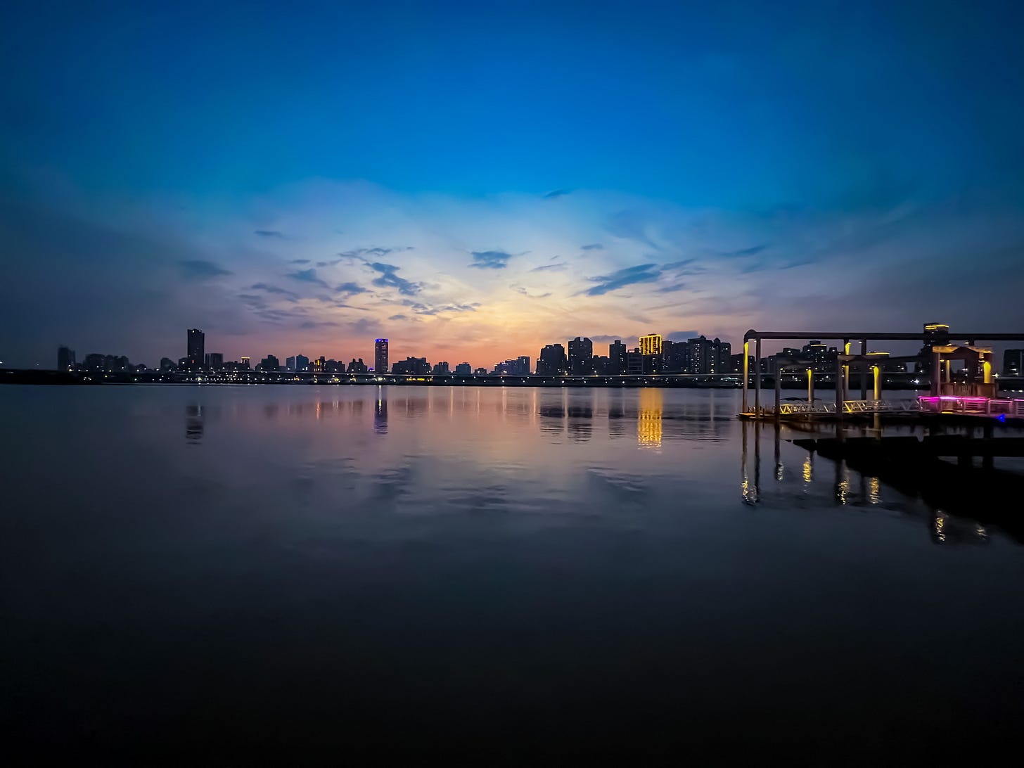 Sunset over the Tamsui river. A lit-up pier juts out into the river, and the skyline of New Taipei visible against the pink sky on the opposite bank