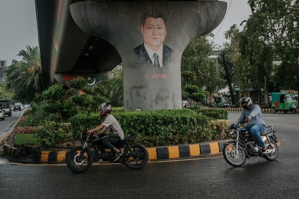 Two people on motorcycles pass a concrete pillar painted with the face of Xi Jinping.