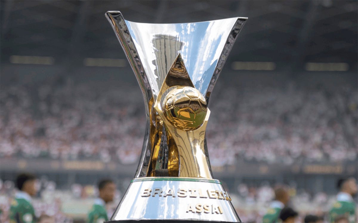 Troféu do Campeonato Brasileiro Série A, no centro da imagem. A taça é prateada, com um detalhe dourado em forma de bola de futebol no centro. Na base, lê-se a inscrição "BRASILEIRÃO ASSAÍ". Ao fundo, desfocado, aparecem arquibancadas lotadas de torcedores Troféu do Campeonato Brasileiro Série A, no centro da imagem. A taça é prateada, com um detalhe dourado em forma de bola de futebol no centro. Na base, lê-se a inscrição "BRASILEIRÃO ASSAÍ". Ao fundo, desfocado, aparecem arquibancadas lotadas de torcedores