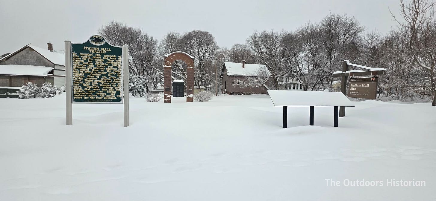 The red sandstone archway of the Italian Hall memorial site in Calumet, Michigan, during winter. Snow covers the ground around the memorial plaques and signs, centering the lone archway against a quiet, wintry backdrop.