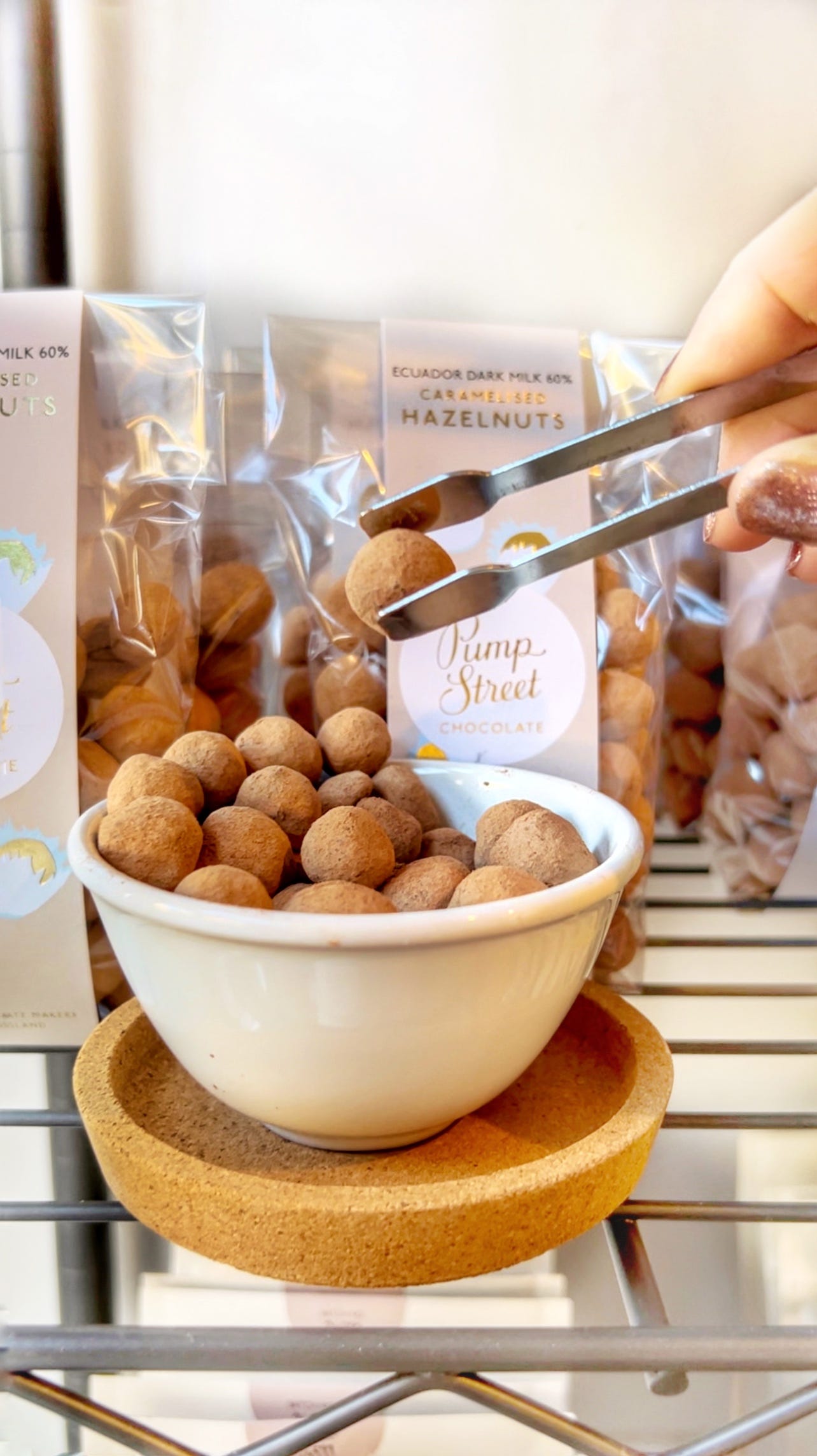 Bowl of Pump Street chocolate-covered hazelnuts with bags of them behind. One is being lifted by giant tweezers. 