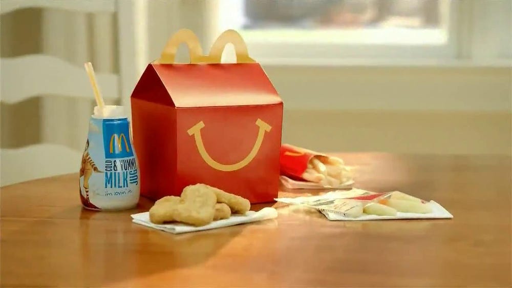 Screenshot of a McDonald's video advertisement, featuring a Happy Meal box, a milk bottle with a straw, several chicken nuggets, French fries, and a few apple slices.