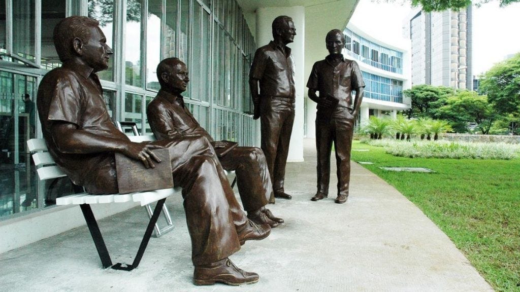 #descrevemospravocê  A imagem mostra um conjunto de esculturas em bronze representando quatro homens em tamanho real, dispostos em frente a um edifício moderno com fachada envidraçada. Duas das figuras estão sentadas em um banco branco, enquanto as outras duas estão de pé, como se participassem de uma conversa informal. As esculturas estão posicionadas sobre uma calçada de concreto, ao lado de uma área gramada com árvores, arbustos e prédios ao fundo.  As figuras têm trajes simples, com camisas de manga curta, calças e sapatos, e seguram livros nas mãos, sugerindo que são intelectuais, escritores ou cientistas. A cena transmite uma atmosfera de diálogo, troca de ideias e valorização do conhecimento.  O edifício ao fundo é curvo, com vários andares e amplas janelas de vidro. O ambiente é arborizado e urbano, com elementos de arquitetura moderna.  Essa instalação artística é conhecida como o "Encontro Marcado", em homenagem aos escritores mineiros Fernando Sabino, Hélio Pellegrino, Otto Lara Resende e Paulo Mendes Campos, e está localizada em frente à Biblioteca Pública Estadual de Minas Gerais, em Belo Horizonte.