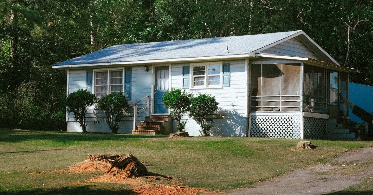 A small single-story white clapboard house with blue shutters, brick front steps and a screened side porch, on a plain lawn with fresh tree stumps in front