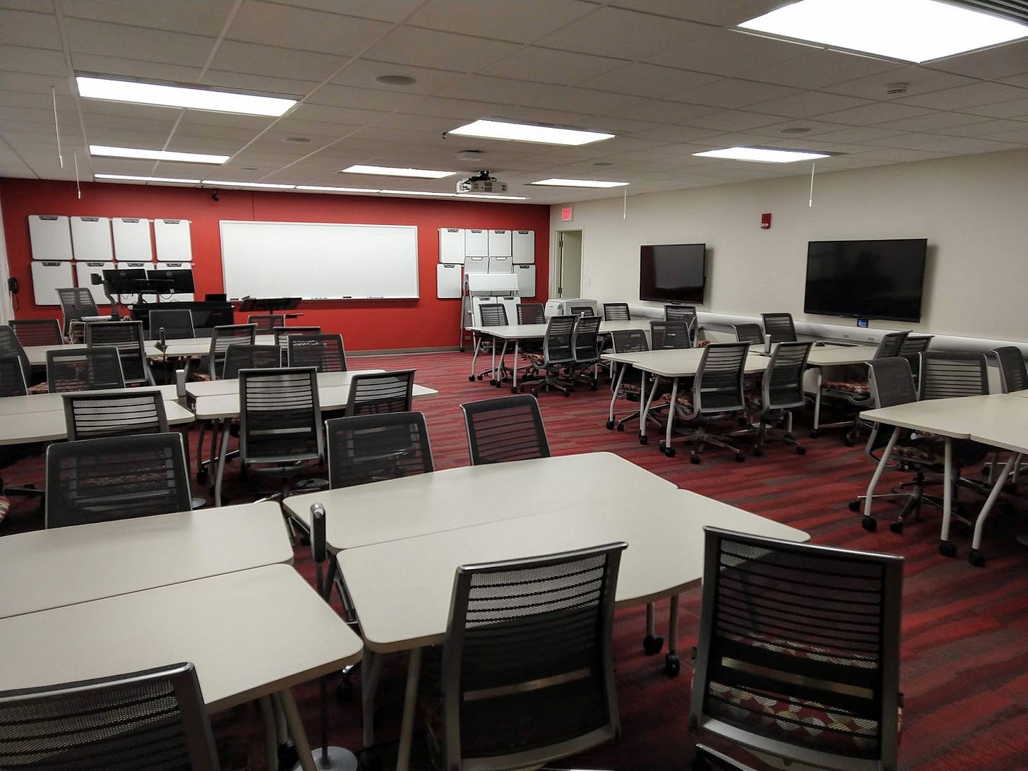TEAL classroom with a large whiteboard and several individual whiteboards on one wall. An instructor podium is near the whiteboards. There are six groupings of tables with eight chairs and a wall mounted monitor each.