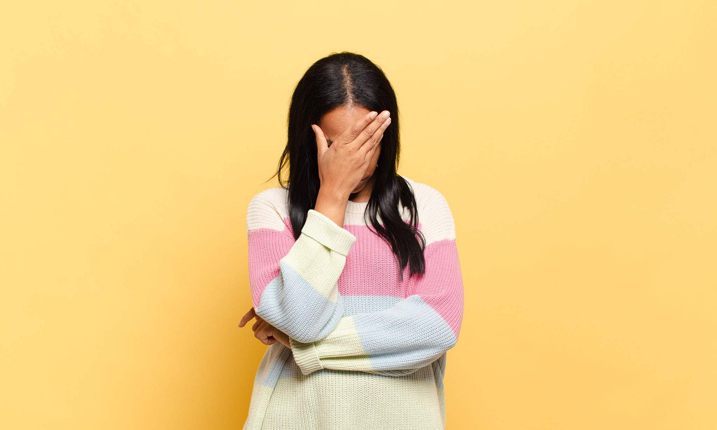 A Black woman with long, straight hair, wearing a pink, blue and white sweater holds her face against her head, covering her eyes. The image is set against a yellow background