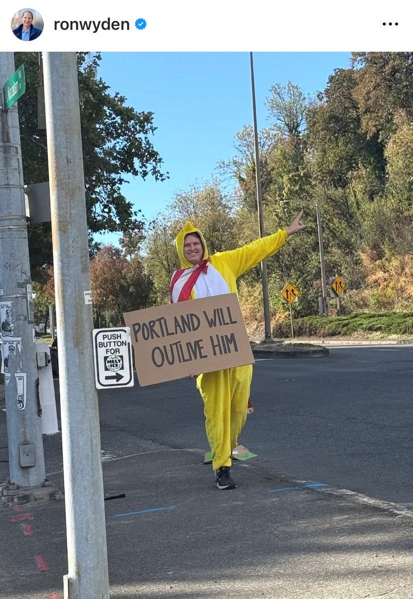 A person in a yellow duck costume holding a sign that reads "PORTLAND WILL OUTLINE HIM." The scene is set on a street with a sidewalk, traffic signs, and trees in the background. Ron Wyden is visible in the top left corner of the image, indicating his presence or association.
