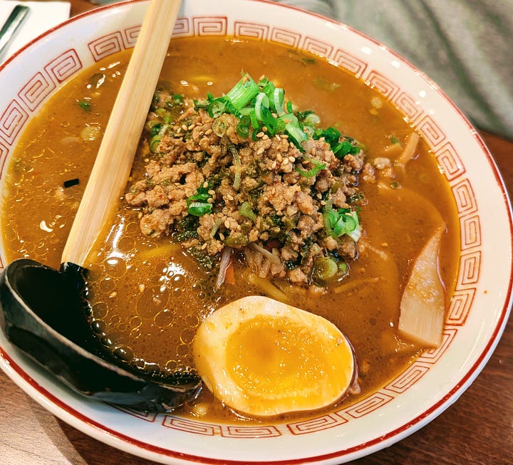 Two ramens. On the left featured noodles, bamboo, green onions, egg, and pork with a long spoon. On the right, a deeper brown/red broth with minced meat. 