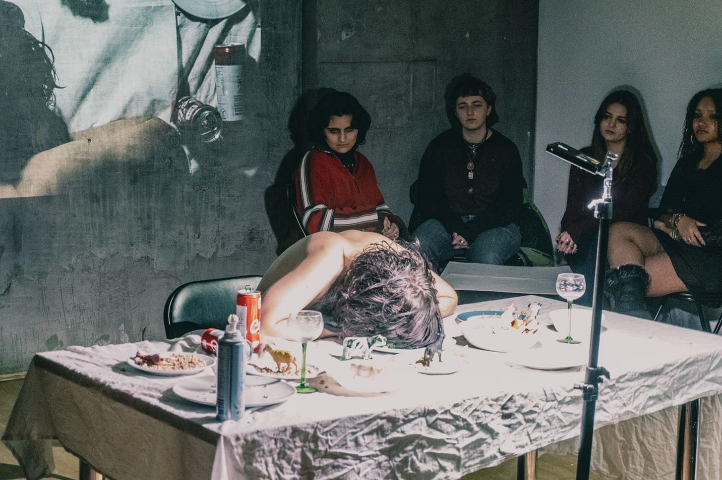 Performance still: A shirtless person with curly hair sits slumped over a table covered in disheveled plates of food, small animal figurines, and a can of whipped cream. A bright light illuminates the table, contrasting with the dark, industrial-looking room. Four audience members watch intently, their faces partially shadowed. A projection on the wall behind them displays a close-up of a similar dining scene.