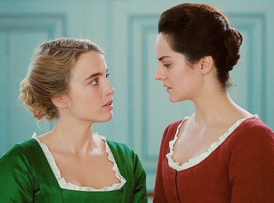 gif: marianne e heloise se olham. marianne usa vestido vermelho e heloise usa um vestido verde