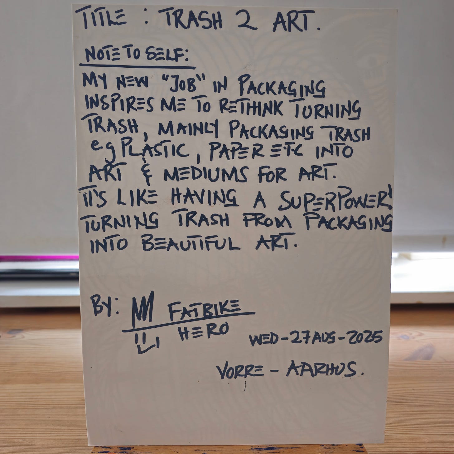 Alt Text: The back of an artwork card containing handwritten black text on a white background. It includes the title "TRASH 2 ART," a note about turning packaging trash into art, the artist's signature "M Fatbike Hero," and the date "Wed-27 Aug-2025."