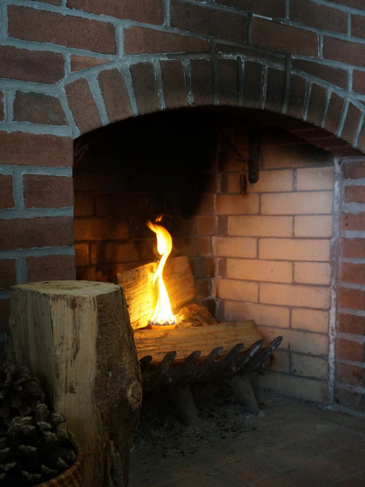 A lit hearth fire glowing inside a brick fireplace, paired with the full Snow Moon rising through bare winter branches — reflecting Imbolc’s threshold between inner warmth and returning lunar light.