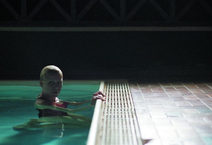 A woman clings to the edge of an indoor swimming pool, half-submerged, staring tensely toward something off-screen. The lighting is dim, adding to the sense of isolation and unease.