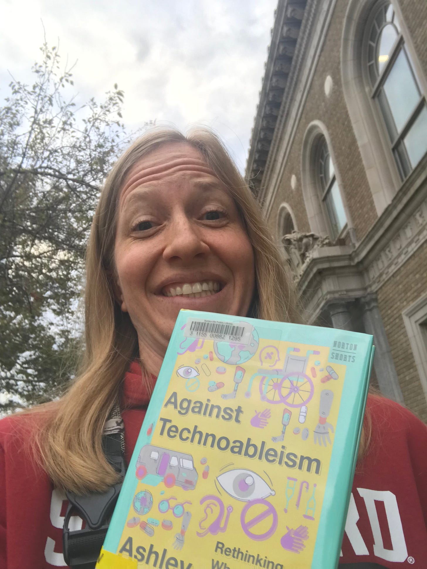 Selfie of Rachel Kolb, a white woman with shoulder-length blonde hair. She is standing in front of a stone library building holding Ashley Shew's Against Technoableism, grinning.
