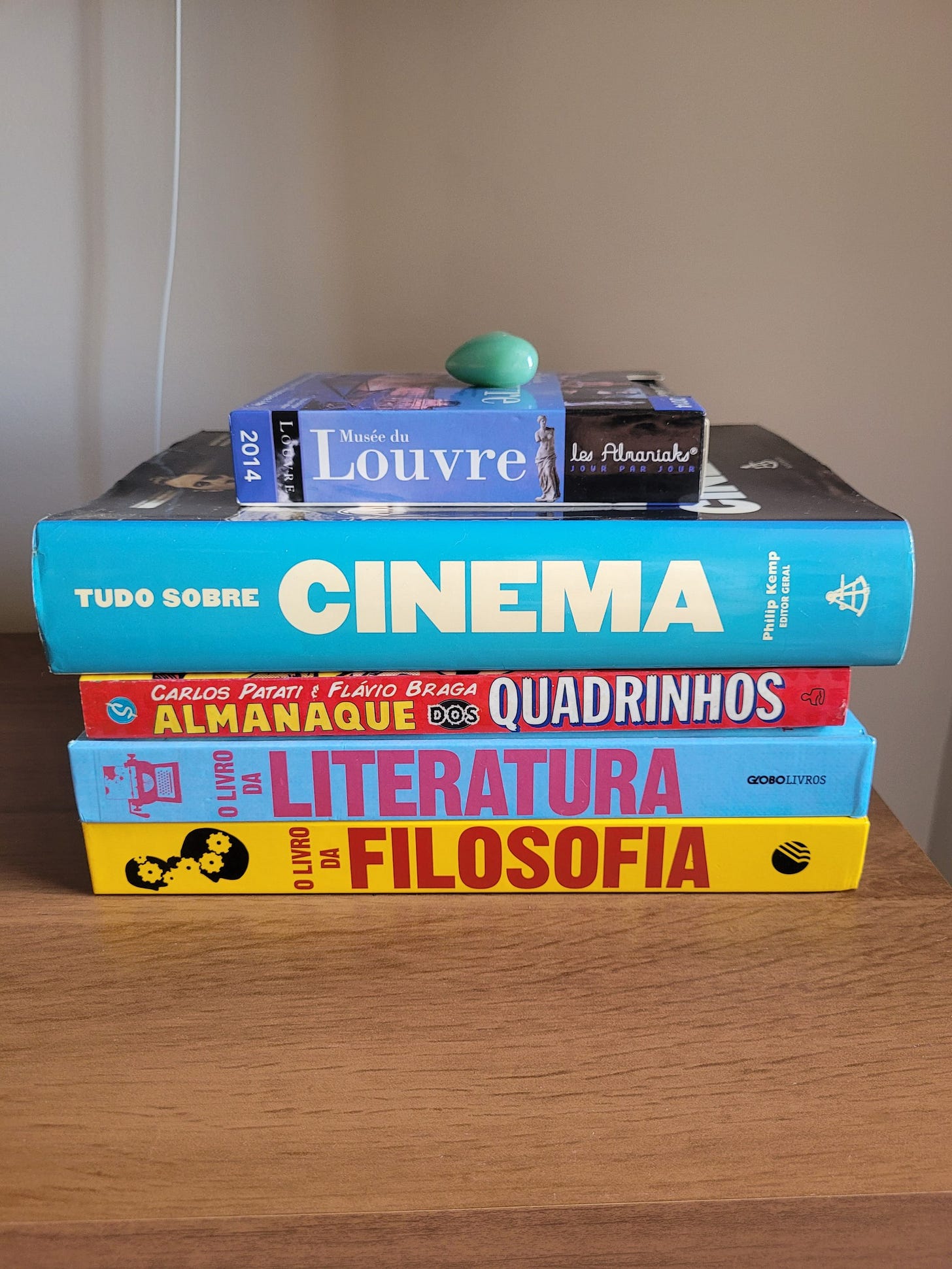 Foto de cinco livros em cima de um rack, deitados, com as lombadas de lado, um por cima do outro. O mais de baixo é amarelo e se chama O Livro da Filosofia. Em seguida, azul claro, O Livro da Literatura. Após este, em vermelho, Almanaque dos Quadrinhos. Depois um muito grosso com lombada azul e texto branco Tudo sobre Cinema. Por fim, um pequeno chamado Musée du Louvre 2014. Acima deste, uma pedrinha verde.