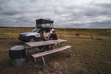 A gallery collage of eight photos from a cross-country road trip. The images show a dark-colored Ford Escape in various scenic American landscapes, including mountains, open plains at sunset, and on the road. The gallery also includes details from the journey and a view from inside the car.