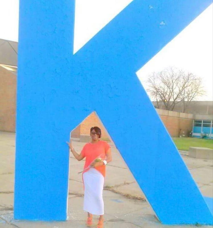 a brown-skinned Black woman with short black hair (long swoop in the front), sunglasses, an orange top, a white maxi skirt, and orange heels stands underneath a giant blue ‘K’. a brown-skinned Black woman with short black hair (long swoop in the front), sunglasses, an orange top, a white maxi skirt, and orange heels stands underneath a giant blue ‘K’.