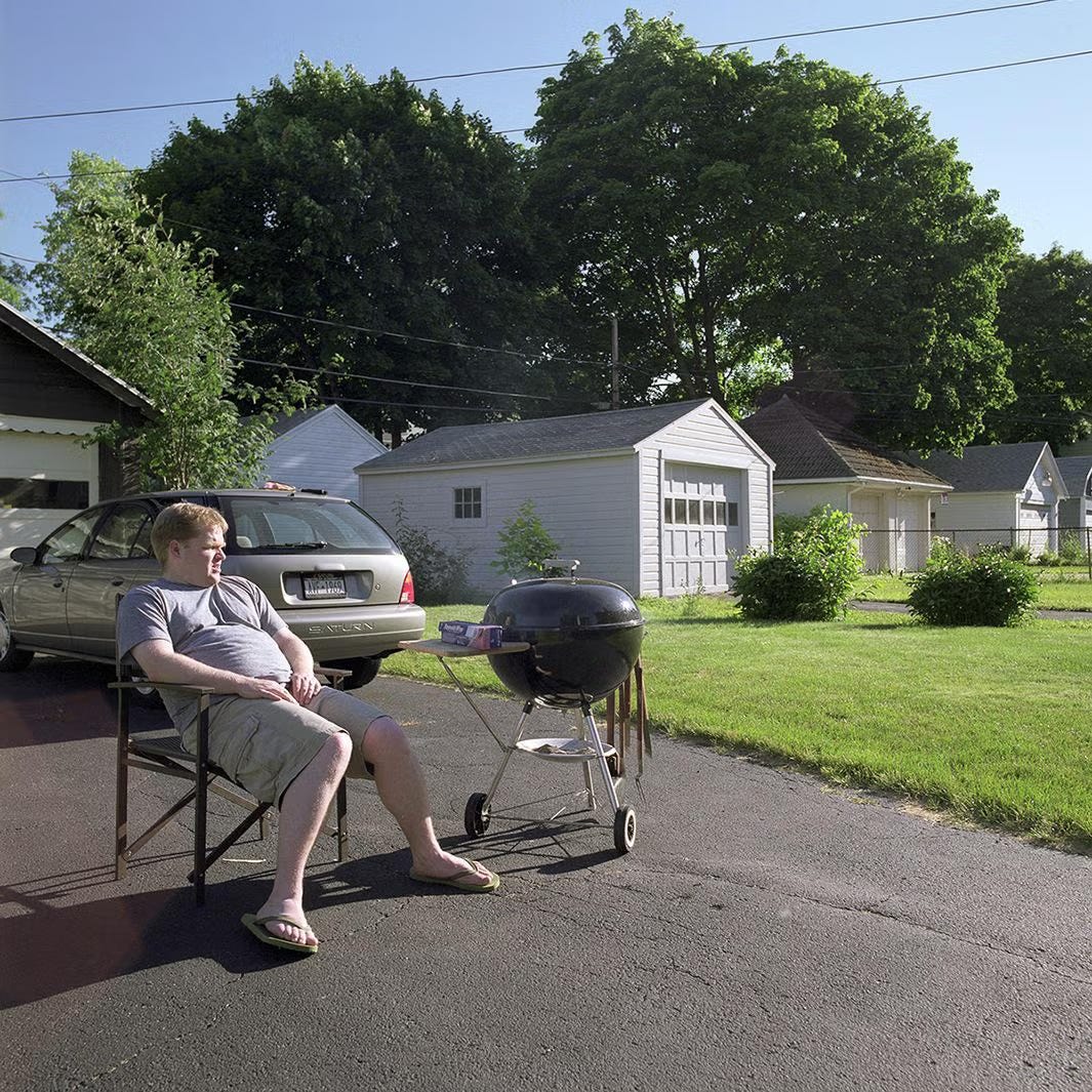 man sitting in lawn chait in driveway beside grill; suburbs around him man sitting in lawn chait in driveway beside grill; suburbs around him