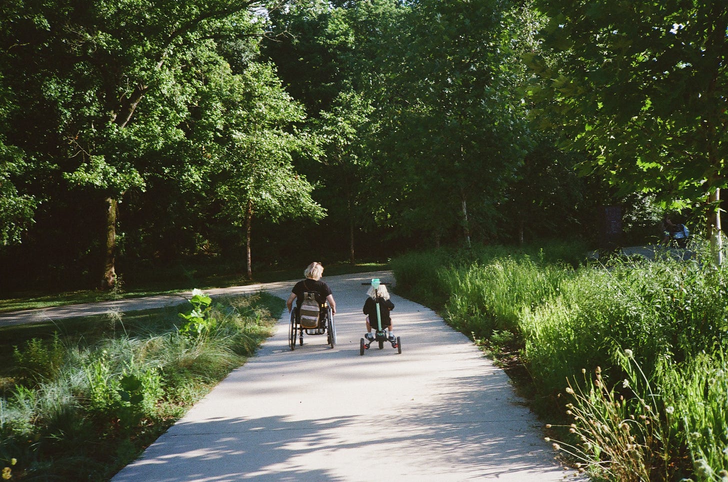 A photo taken from behind of Rebekah and her young son as they zoom down a trail surrounded by green trees and grass. Rebekah sits in wheelchair and she’s looking toward the right at her son who is pedaling on his tricycle. Their bodies look small on the wide, long path.