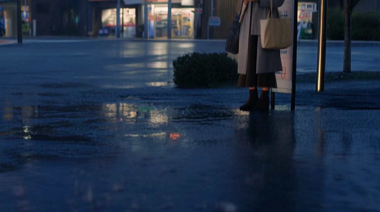 Rain-wet pavement at dusk with shop lights reflected in puddles and a woman waiting at a bus stop