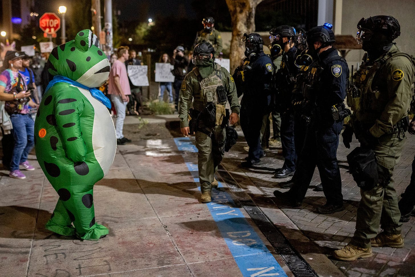 A photo shows a protester in a green inflatable frog costume on the left, facing law enforcement officers in riot gear at night.   