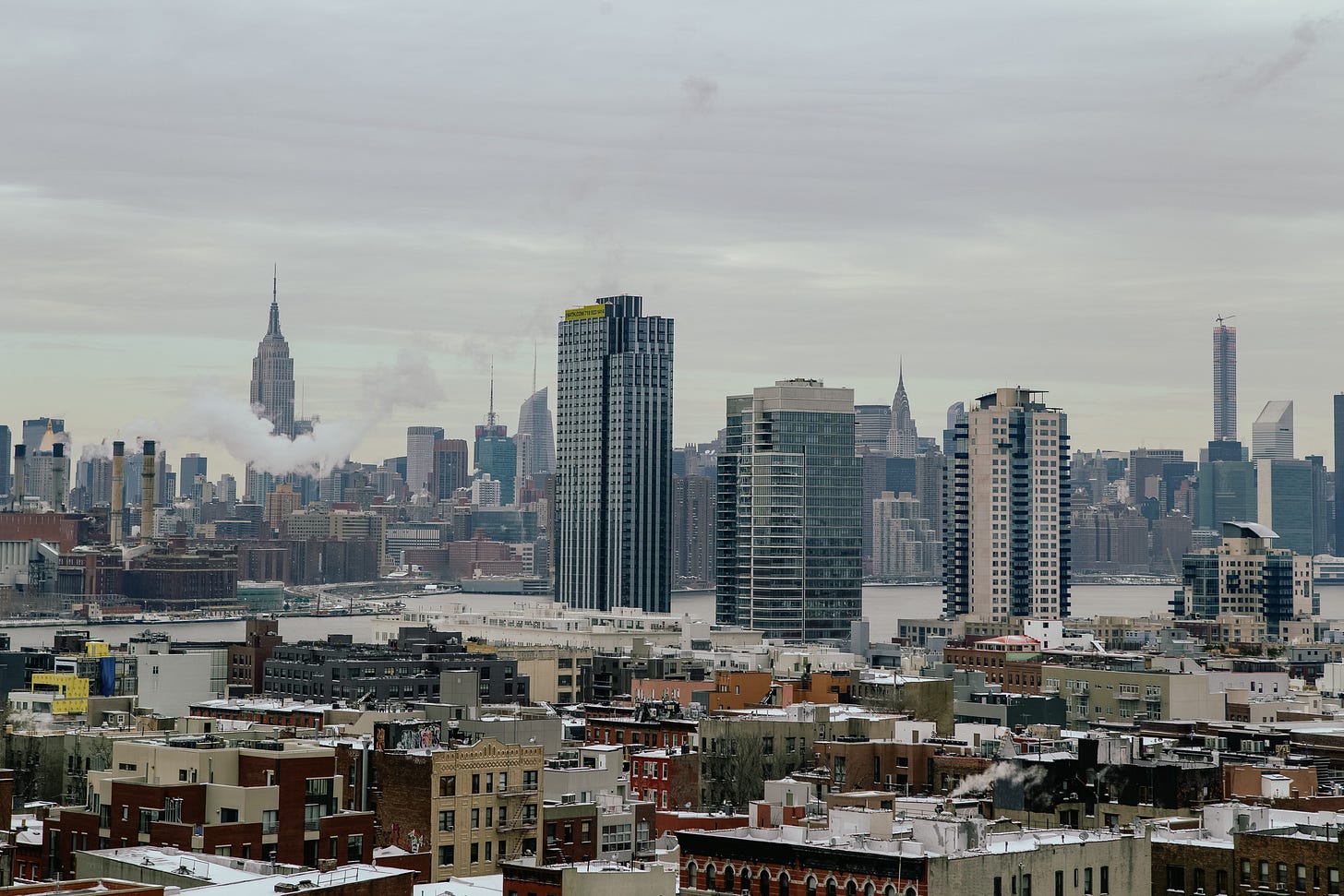 A wide view of the Midtown Manhattan skyline on a cloudy winter day, seen from across the East River. The rooftops in the foreground are dusted with snow. The Empire State Building is prominent on the left side of the skyline, with steam rising from a nearby building.
